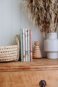 some books are sitting on a dresser next to a basket and vase with dried grass in it