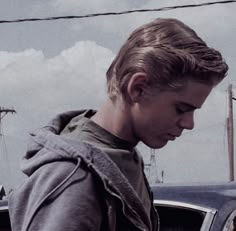 a young boy standing in front of a car looking at his cell phone while wearing a hoodie