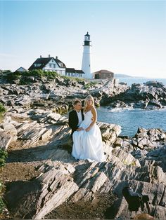 two women sitting on rocks near the ocean and a lighthouse in the background with blue sky