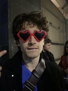 a man with heart shaped glasses on his eyes pointing at the camera while standing in front of a wall