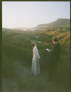 a bride and groom standing in the sand at sunset, reading vows to each other