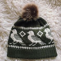 a green and white knitted beanie hat with two birds on it, sitting on a furry surface