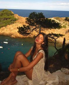 a beautiful young woman sitting on top of a rock next to the ocean with boats in the water