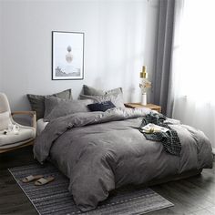 a bed with grey linens and pillows in a white room next to a window