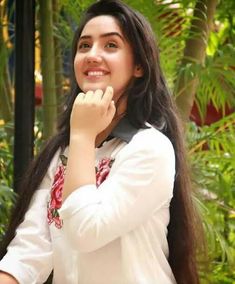 a woman with long hair smiling and holding her hand up to her face in front of some plants