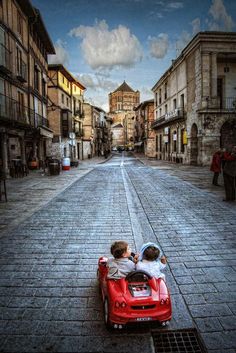 two children are riding in a toy car down the street with buildings on either side