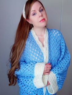 a woman wearing a blue crochet sweater and white knitted headband standing in front of a gray wall