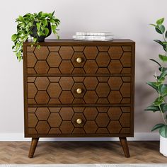 a wooden chest of drawers with plants on top and books on the bottom, in front of a white wall