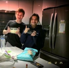 two young men standing in a kitchen holding up their camera's thumb and giving the thumbs up sign