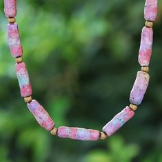 a pink and blue necklace with gold beads on a tree branch in front of some trees