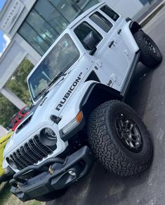 a white jeep is parked in front of a building with large tires on it's wheels