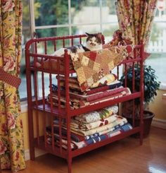 a cat sitting on top of a red shelf next to a window filled with quilts