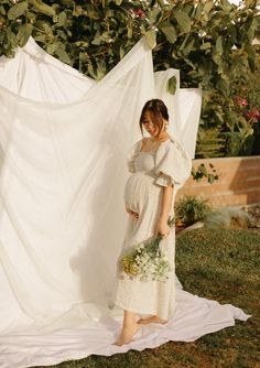 a pregnant woman wearing a white dress standing in front of an unfurnished backdrop