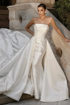 a woman in a wedding dress leaning against a stone wall with her hand on her hip