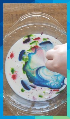 a child's hand touching the bottom of a bowl filled with melted paint on top of a wooden table