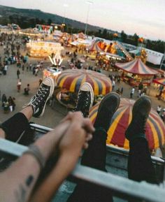 two people sitting on the edge of a carnival looking down at an amusement park filled with rides and fair goers