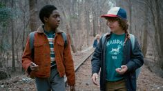 two young men standing in the woods talking to each other and looking at one another