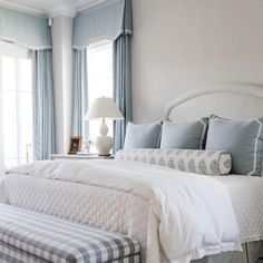 a white bed sitting next to a window with blue curtains and pillows on top of it