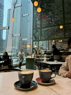 two cups of coffee sit on a table in front of a window with people sitting at tables