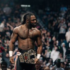 a man with no shirt on standing in front of a crowd holding a wrestling belt