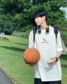 a young man holding a basketball while standing on top of a lush green field next to a park