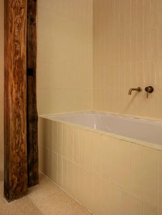 a bath tub sitting next to a wooden pole in a bathroom with beige tiles on the walls