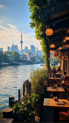 people are sitting at tables near the water in front of some tall buildings and trees