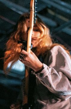 a woman with long red hair holding a guitar in her right hand and looking down at the strings