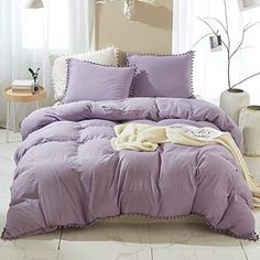 a bed with purple comforter and pillows in a room next to a lamp on a table