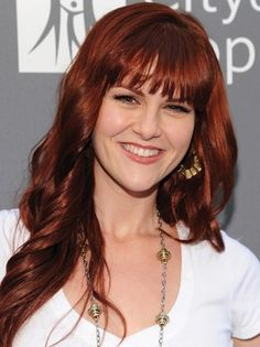 a woman with long red hair wearing a white shirt and gold earrings smiles at the camera