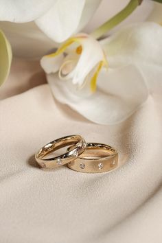 two gold wedding rings sitting next to each other on a white cloth with flowers in the background