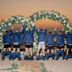 a group of men standing on top of a sandy beach under a white flower covered arch