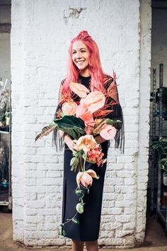 a woman with pink hair standing in front of a white brick wall holding a bouquet of flowers