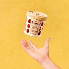 a hand reaching out towards an ice cream container with fruit on it, in front of a yellow background