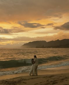 a man and woman standing on top of a beach next to the ocean at sunset