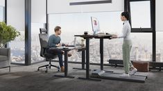 two people sitting at desks in an office with large windows looking out on the city