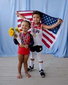 two young children standing next to each other in front of an american flag and holding flowers