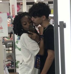 two people standing in front of a store looking at their cell phones and kissing each other