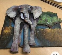 an elephant made out of paper sitting on top of a wooden table next to a painting