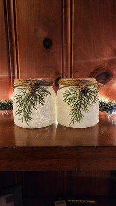 two mason jars decorated with pine cones and evergreen needles are sitting on a wooden table