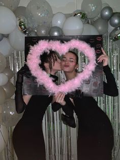 two women holding up a pink heart shaped photo frame in front of balloons and streamers