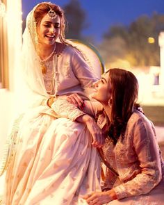 two women in wedding dresses sitting next to each other on a bench and one is touching the woman's face