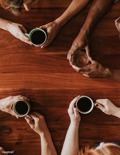 four people holding cups of coffee on top of a wooden table with their hands in the air