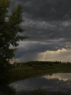 storm clouds loom over a small pond in the middle of a grassy area with trees