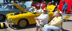 two people sitting in chairs next to parked cars with open hoods and sunflowers on their heads