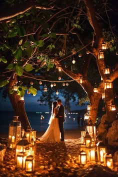 a bride and groom standing under a tree surrounded by lit up lanterns on the beach