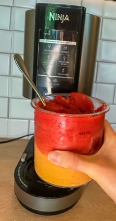 a hand holding a cup filled with fruit on top of a counter next to a blender