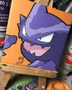 a painting of a purple cat on an easel next to cans of sodas