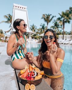 two women sitting next to each other in front of a swimming pool with fruit on the table