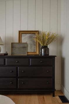 a black dresser with flowers and pictures on it in a white walled room next to a bed